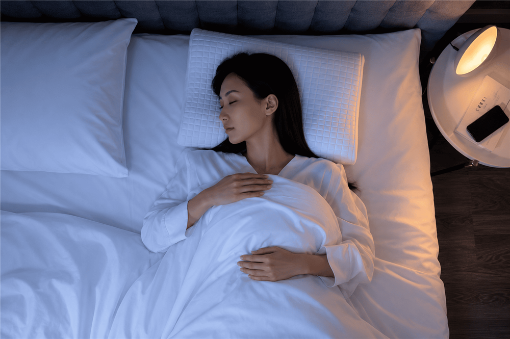 Woman sleeping on an ergonomic pillow in a dimly lit bedroom with a bedside lamp and phone nearby.